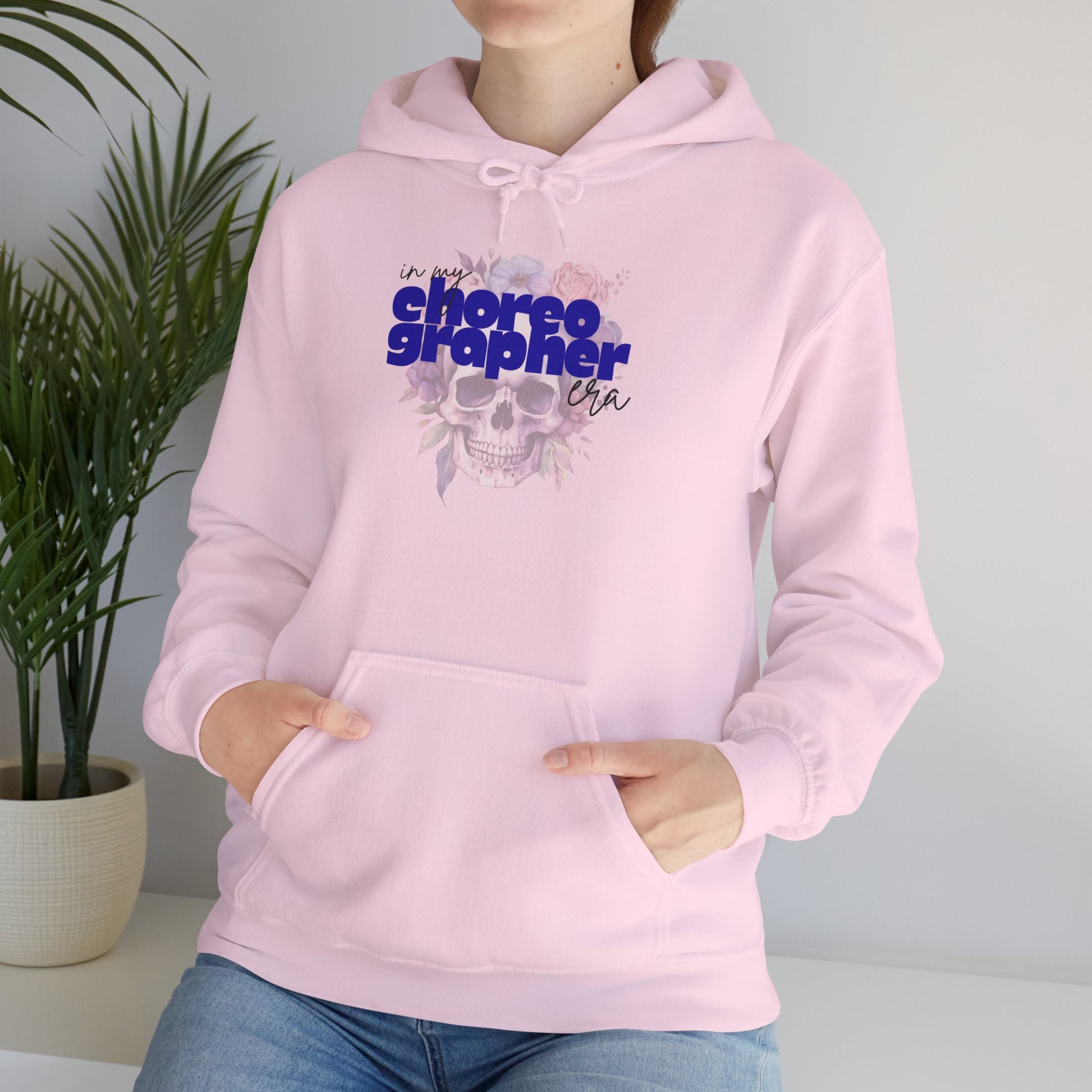 Person wearing a pink hoodie with 'choreographer' text and skull design, standing next to a plant.