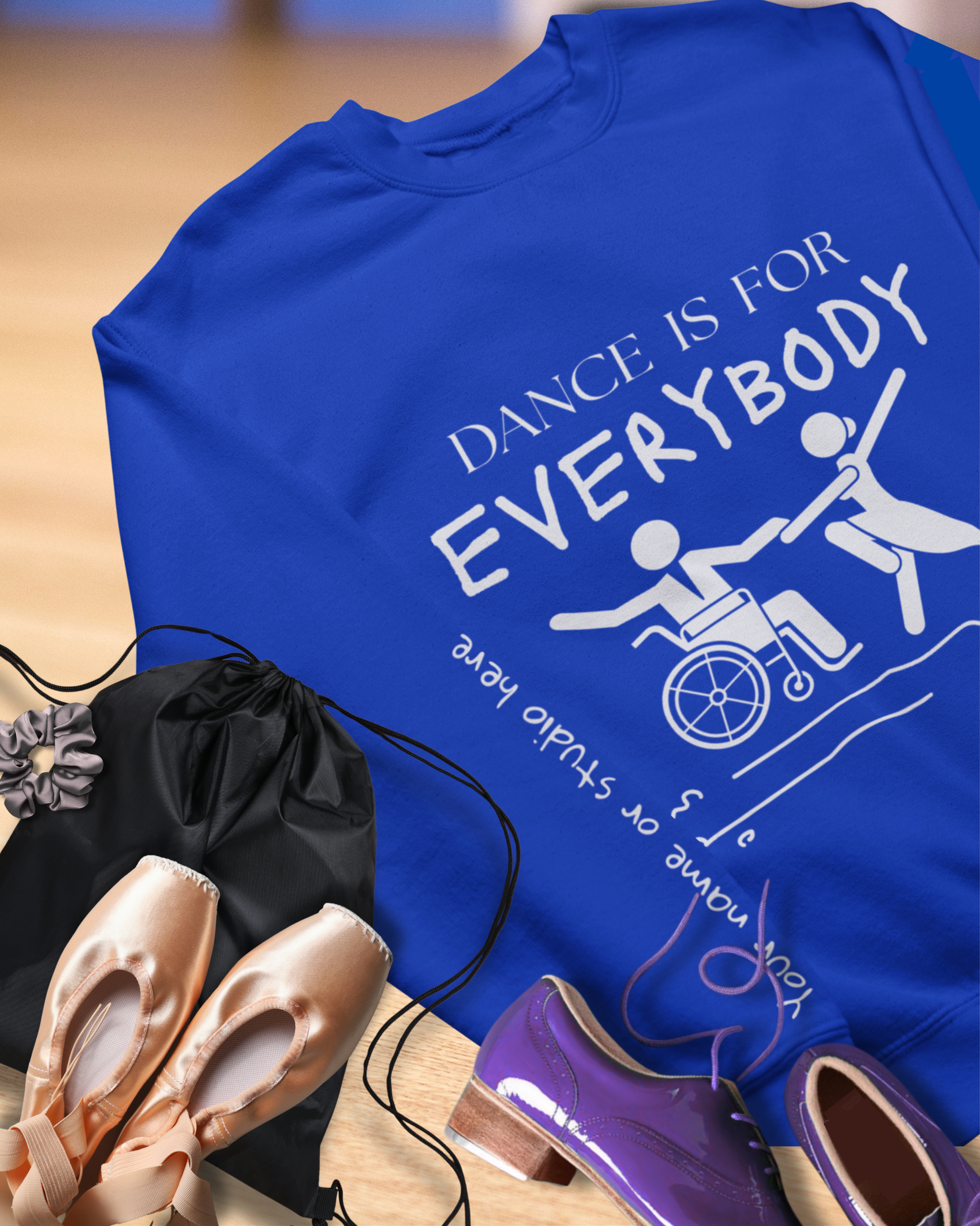 Blue t-shirt with 'Dance is for Everybody' text and wheelchair icon, next to ballet shoes and a black bag.
