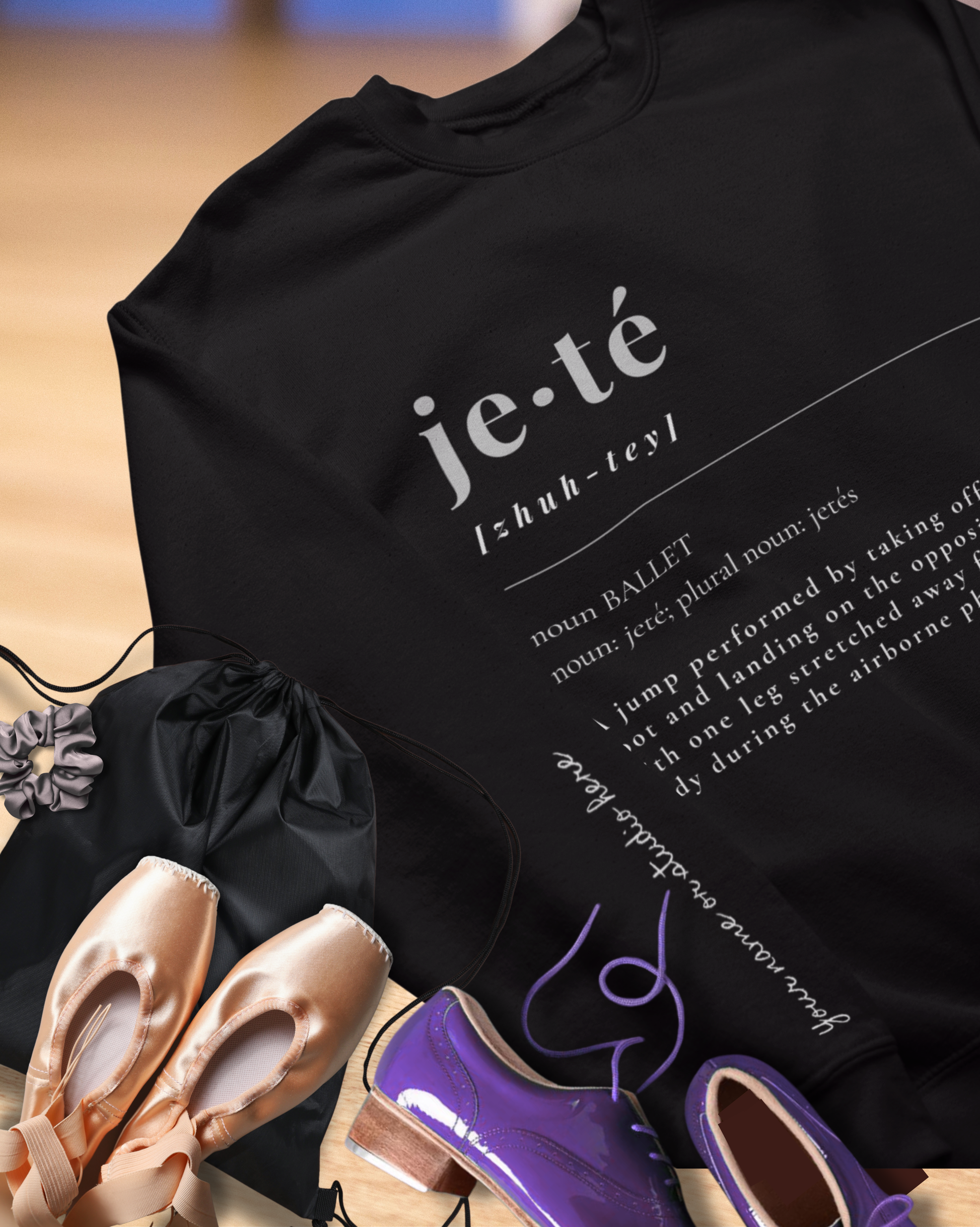 Black hoodie with 'je-te' text and definitions, placed on a wooden surface with ballet shoes around.